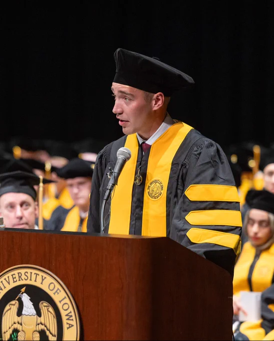 A man in academic regalia stands at a podium adorned with the University of Iowa seal, delivering a speech at a commencement ceremony. He's wearing a black cap and gown with gold accents, matching the seated graduates in the background, who also wear similar robes and caps. A microphone is positioned in front of him as he addresses the audience.