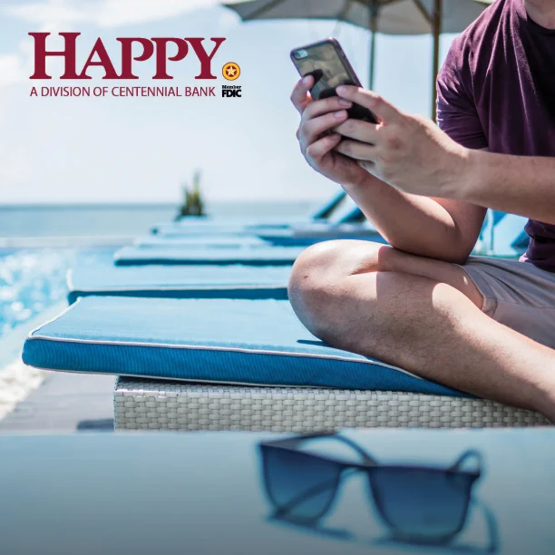 Person sitting cross-legged on a poolside lounge chair using a smartphone under a sun umbrella on a sunny day.
