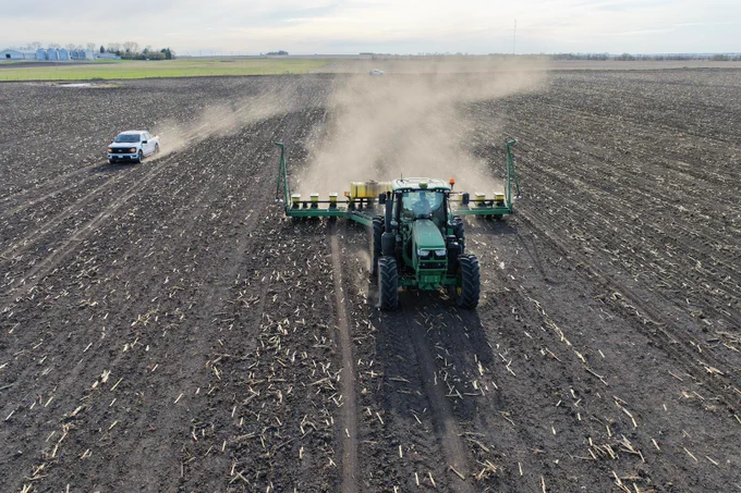 A green John Deere tractor is pulling a large planter across a vast, freshly plowed field, with dust rising behind it. A pickup truck drives parallel to the tractor. The horizon shows a flat landscape under a partly cloudy sky.