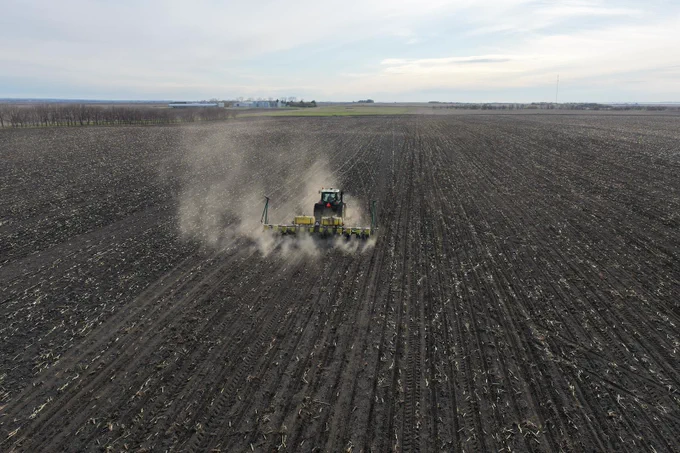 A tractor plows a vast field, stirring up dust. The landscape stretches into the distance under a cloudy sky. Sparse trees border the field on the left, and some distant structures are visible on the horizon.