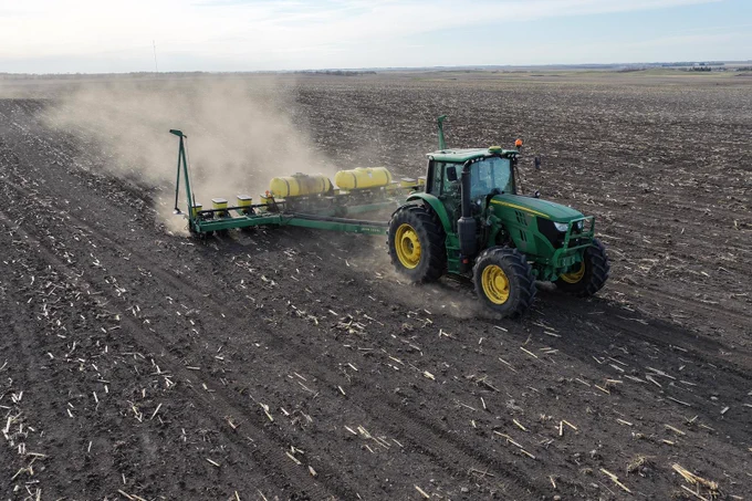A green John Deere tractor is pulling a large planter through a vast, plowed field, creating clouds of dust. The sky is overcast, and the landscape is expansive and rural.
