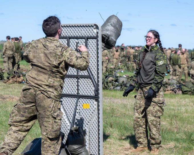 Two soldiers in camouflage engage in a training exercise using sandbags on a grassy field with others in the background.