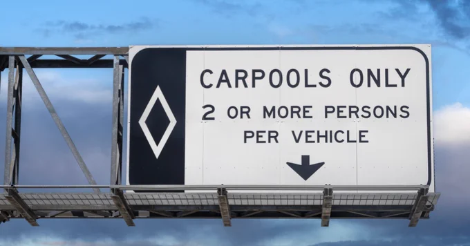 A highway sign against a cloudy sky reads: "Carpools Only. 2 or more persons per vehicle," with an arrow pointing downward and a diamond symbol on the left.