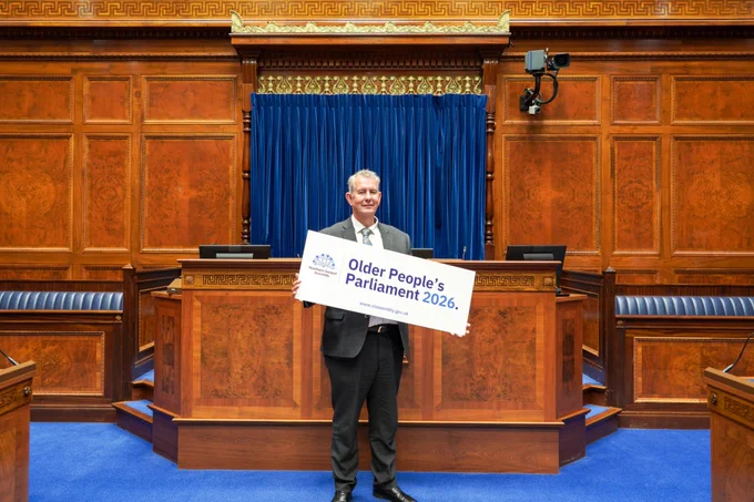 A man in a dark grey suit jacket, white shirt, and patterned tie is holding a large white sign that reads "Older People's Parliament 2026" in blue text, with the Northern Ireland Assembly logo of blue flax flowers on the left. He is standing in the centre of a wood-panelled parliament chamber with blue carpets and a blue curtain. 