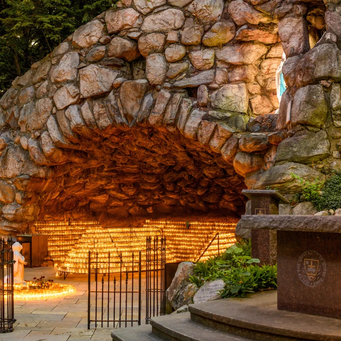 A stone grotto illuminated by numerous candles arranged on shelving within the arch. A statue of a religious figure is visible to the left. The setting is serene and suggests a place of worship or reflection.