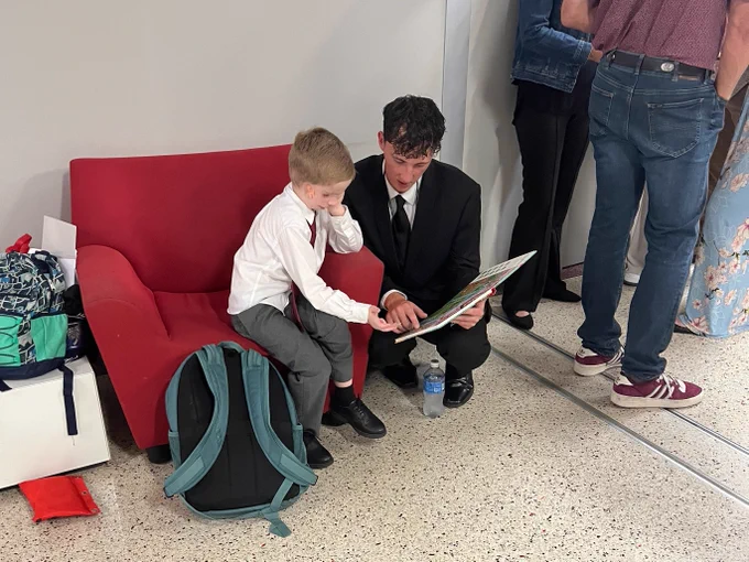 A person in formal attire sits on the arm of a red chair, reading a book with a child seated on the chair. The child is dressed in a white shirt and gray pants, with a backpack nearby. Other individuals are partially visible in the background.