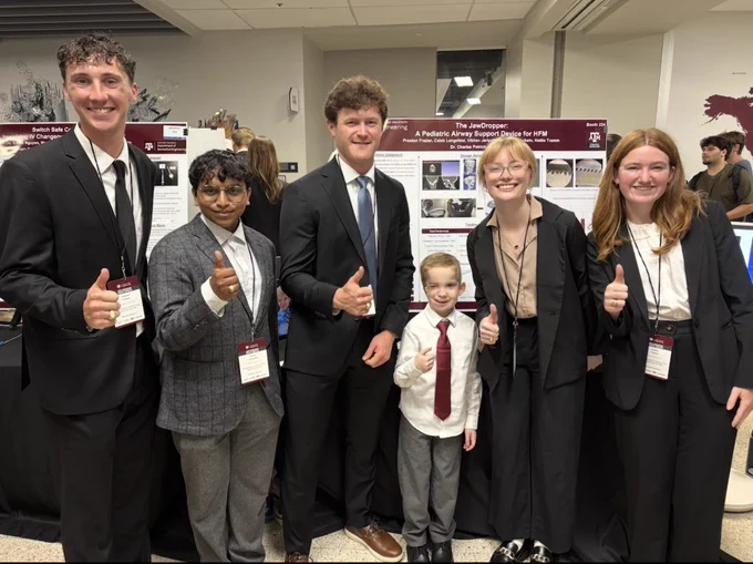 A group of six people stand in front of informational posters at a presentation event. Five adults are dressed in formal attire, and a child is also wearing a shirt and tie. They are giving a thumbs-up gesture. The atmosphere is professional and friendly.