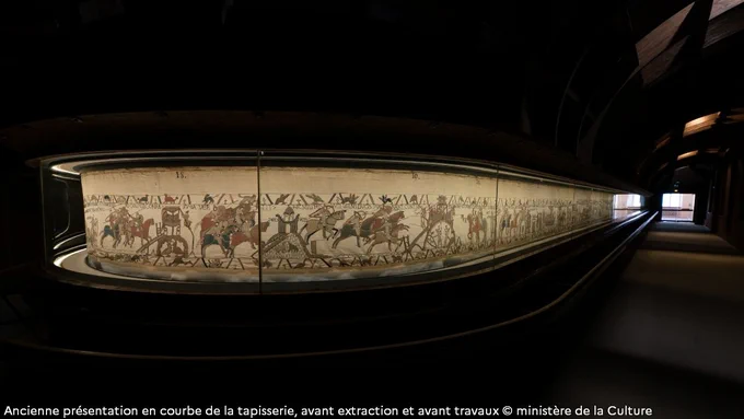 Ancienne présentation en courbe de la tapisserie, avant extraction et avant travaux © ministère de la Culture
Dans la pénombre, la tapisserie de Bayeux est présentée dans une vitrine avec des courbes.