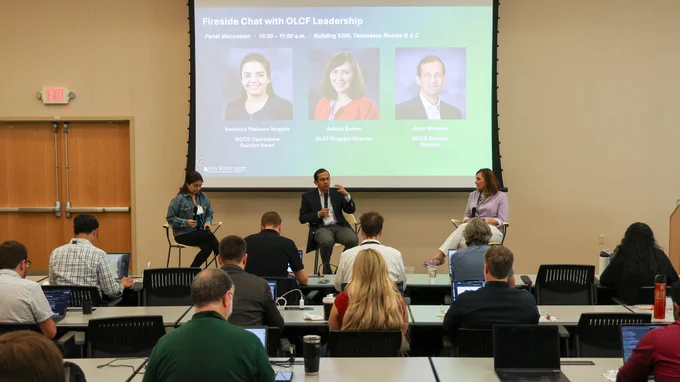Attendees seated at tables with laptops listen to a fireside chat featuring Verónica Melesse Vergara, Arjun Shankar and Ashley Barker during the Oak Ridge Leadership Computing Facility User Summit. A large screen behind the speakers displays their names and leadership roles.