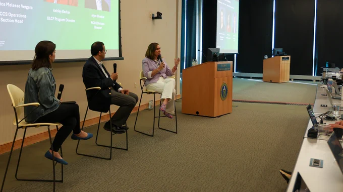 Ashley Barker speaks into a handheld microphone while participating in a fireside chat with Oak Ridge Leadership Computing Facility leadership. Verónica Melesse Vergara and Arjun Shankar sit nearby as attendees listen from the audience.