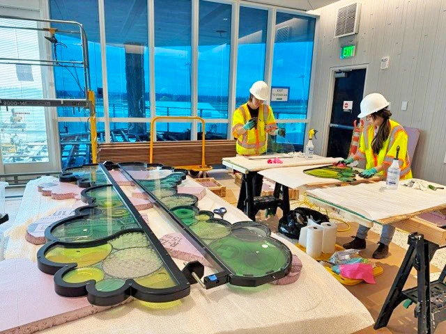 Two people in hard hats working on large, colorful panels in a well-lit room with big windows.