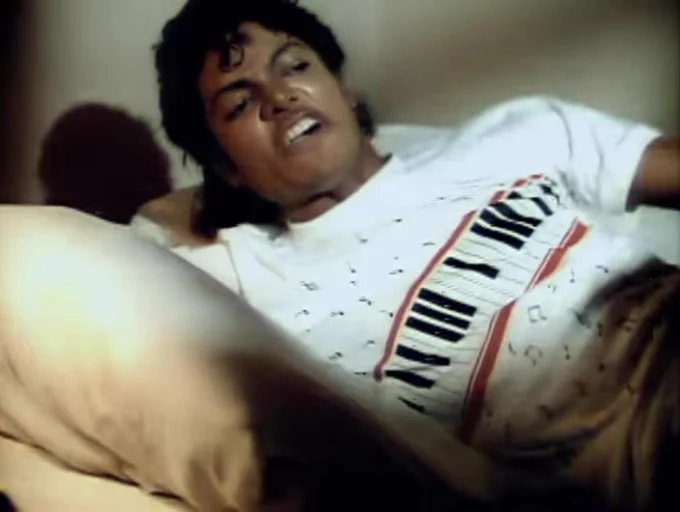 Screenshot aus MJ's Beat It-Video. Er liegt auf einem Bett und trägt ein weißes Shirt. Diagonal über der Brust verläuft eine Klaviatur, doch die Tasten sind falsch dargestellt. Also mal einzelne schwarze Tasten, dann mal drei nebeneinander, dann wieder zu viele weiße Tasten bis zur nächsten schwarzen Taste usw.