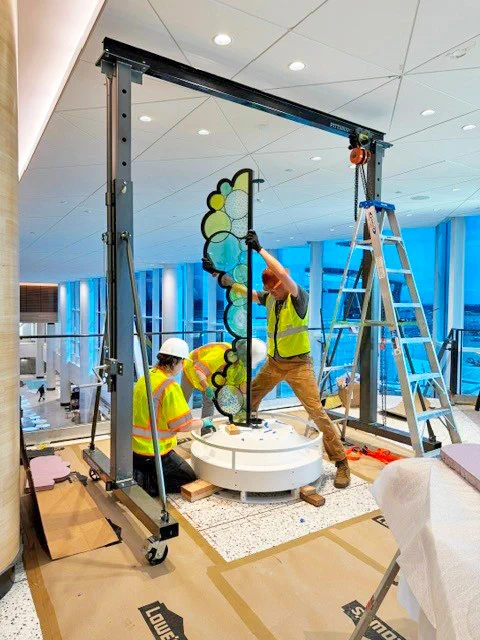 Workers installing a multicolored glass panel inside a large room with blue windows.