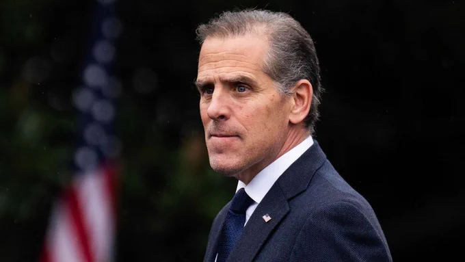 A photo shows Hunter Biden wearing a suit and dark blue tie with a white collared shirt, as well as an American flag pin, with an out-of-focus background displaying an American flag.