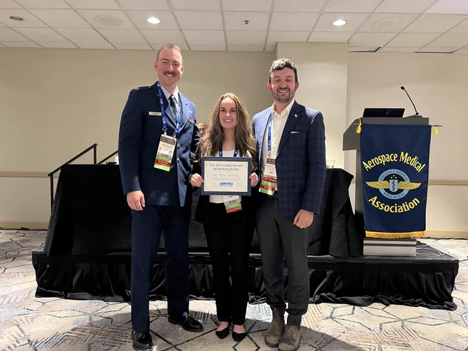 Clare McNerlin stands between two attendees while holding the Anita Mantri, PhD Memorial Award certificate beside an Aerospace Medical Association podium.