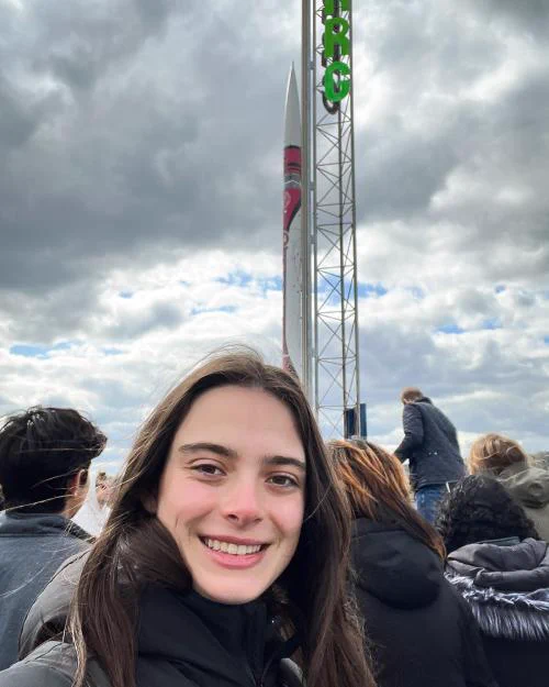 Gabriella Best smiling in the foreground with a small rocket on a launch scaffolding behind. 