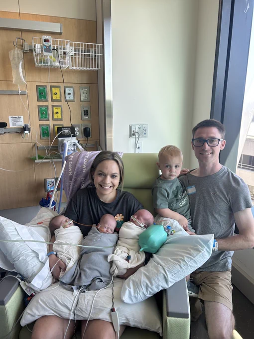 A smiling woman is seated in a hospital room, cradling three swaddled infants with medical tubes. A man stands beside her holding a toddler. Medical equipment is seen on the wall behind them, and sunlight streams in through a large window.