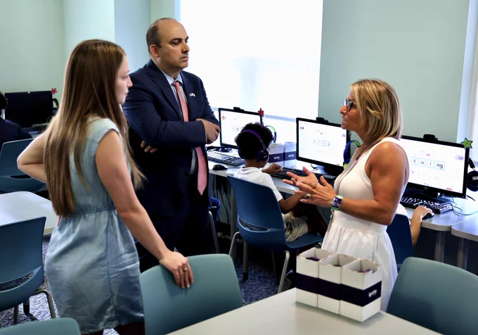 Secretary Zrike in a shirt and tie speaks with two literacy instructors in a library room. Students wear headsets and work at computers behind them.