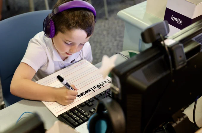 A young student wearing purple headphones participates in a literacy tutoring session at a computer workstation. The student writes on a lined practice sheet while using a keyboard and monitor as part of a structured reading intervention program.
