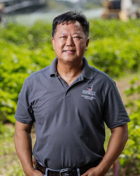 Person standing outdoors in front of a blurred green field. They are wearing a gray polo shirt with the University of Georgia logo visible.