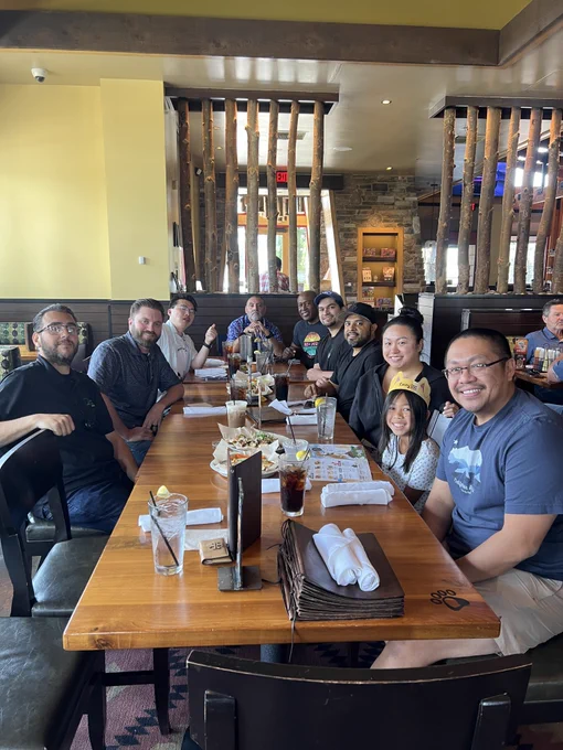 A large, diverse group of coworkers or friends are smiling and posing together around a long wooden dining table inside a restaurant. Food, drinks, and menus are scattered across the table. In the foreground on the right, a man wearing a California Republic t-shirt smiles next to a young girl wearing a paper crown. The restaurant interior features warm lighting, stone accents, and vertical log dividers in the background.