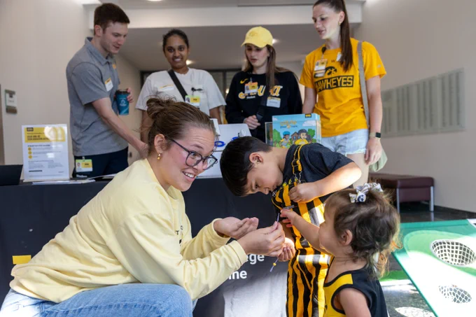 A woman in a yellow sweater crouches to interact with two young children. The boy, wearing a black and yellow striped outfit, is engrossed in a game, while a younger girl in a similar outfit looks on. Behind them, four adults stand, two of them in matching yellow and black 'Hawkeyes' attire, smiling as they oversee a table set up with informational flyers and a game. The environment suggests an engaging community or educational event.