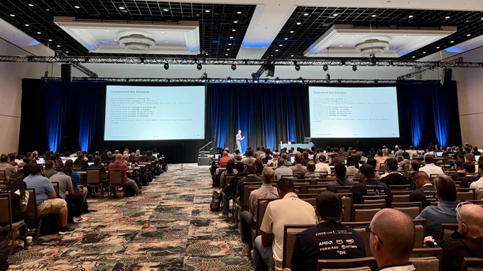 A man presents on stage at a large conference, facing a seated audience. Two screens display \"Implement the Solution\" and network configuration code. Many audience members wear shirts with \"CISCO Live! Las Vegas, NV May 31-June 4, 2025\" and logos like AMD, AWS, & NTT DATA.