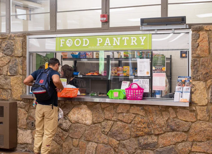 A person stands at a stone counter in front of the UTEP Food Pantry window, selecting items from orange and pink baskets. The sign above reads "Food Pantry," and shelves with various food products are visible behind the window. Flyers and notices are posted on the counter.
