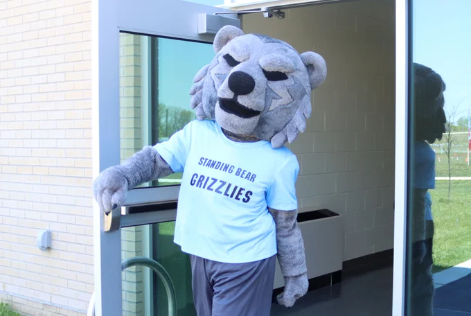 The Standing Bear mascot walks out a door, dressed in a blue t-shirt