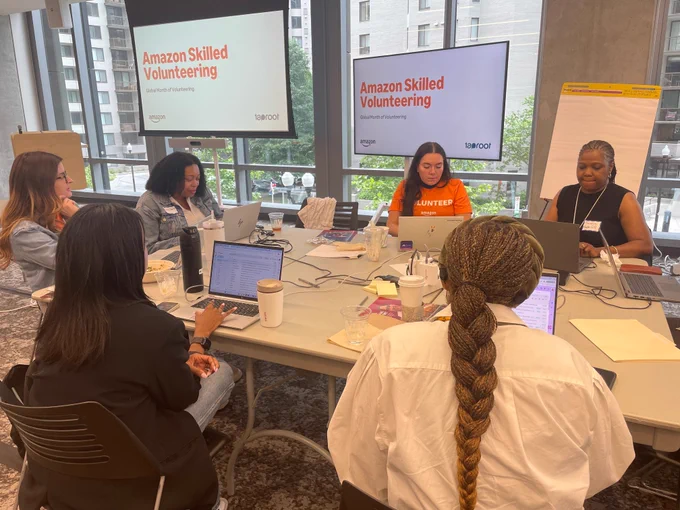 Six people sit around a table with laptops in front of them. Two screens in the background read "Amazon Skilled Volunteering" Taproot and Amazon logos are visible.