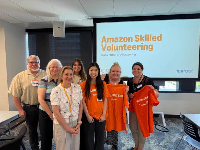 Seven people stand together in front of a screen that reads "Amazon Skilled Volunteer Global Month of Volunteering" Taproot and Amazon logos are visible.