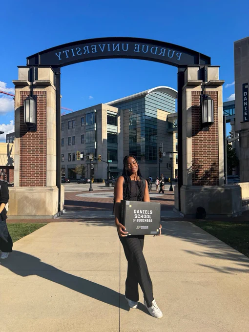 A person stands in front of the Purdue University archway, holding a box that reads "Daniels School of Business." The background features a modern academic building and a clear blue sky.