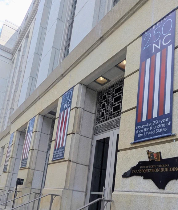 Exterior of the State of North Carolina Transportation Building with banners reading "America 250 NC," noting 250 years since the founding of the United States. The building is constructed from stone and features a decorative entrance.