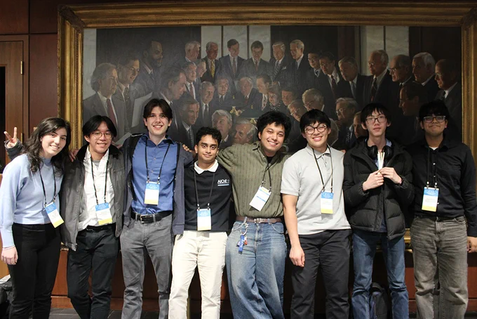 A group of smiling individuals stands in front of a mural depicting past U.S. presidents, including John F. Kennedy. They are casually dressed, wearing conference lanyards, and stand close together inside what appears to be a formal setting.