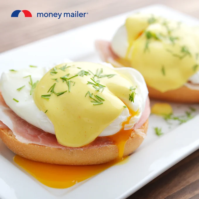Close-up of two eggs Benedict on toasted English muffins with ham, topped with creamy hollandaise sauce and sprinkled with fresh herbs.