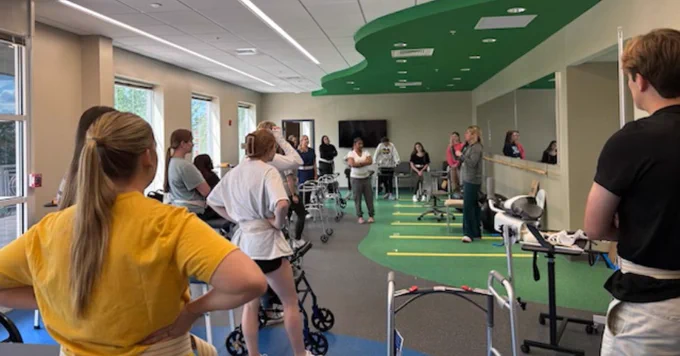 A group of people are in a physical therapy room, equipped with walkers and therapy equipment. The room has green and blue flooring, large windows, and a mirror on one wall. A person at the front is instructing. There is a wall-mounted TV screen in the background.