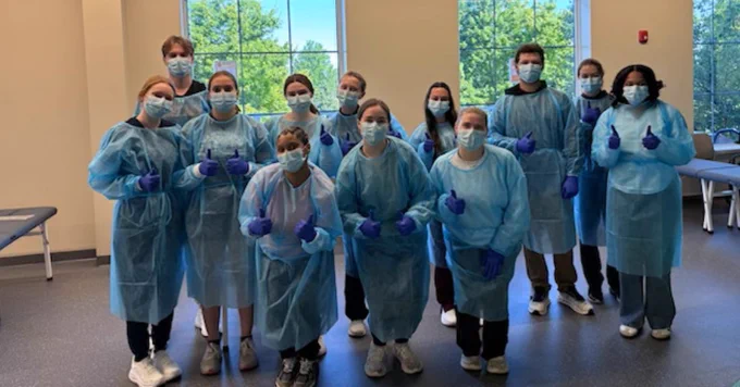 A group of people wearing protective gowns, masks, and gloves stand indoors, facing the camera with thumbs up. Large windows in the background show a sunny view with green trees.