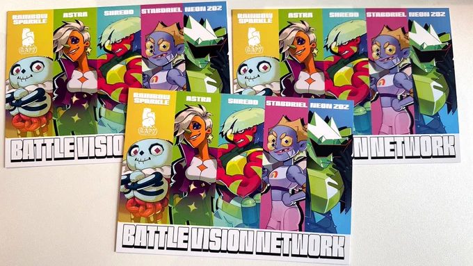 Three postcards are laid out on a table. The postcard features 5 Captains from Battle Vision, each in their own column. The columns are different colours: yellow, green, teal, magenta, and blue respectively. The names of the characters are on the top: Rainbow Sparkle, Astra, Shredd, Stabdriel, and Neon Zoz respectively. It says Battle Vision Network at the bottom of the postcard.