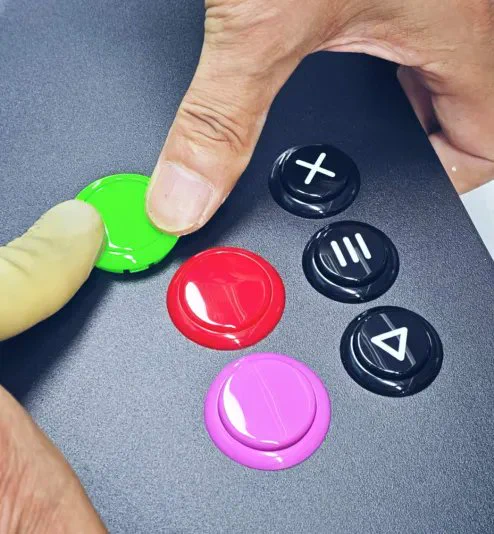 Two fingers pressing colorful arcade buttons on a black surface, including green, red, purple, and black with symbols.