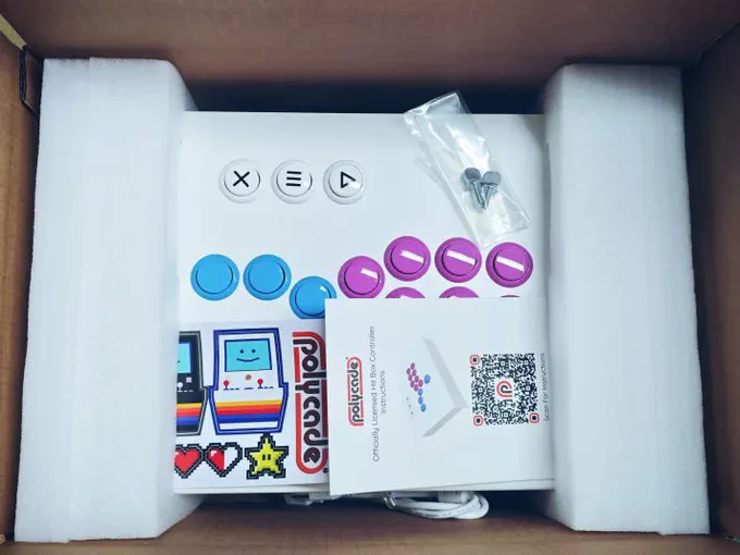 Open packaging showing colorful retro Polycade stickers, instruction manual, screws, and foam padding inside of the controller box.