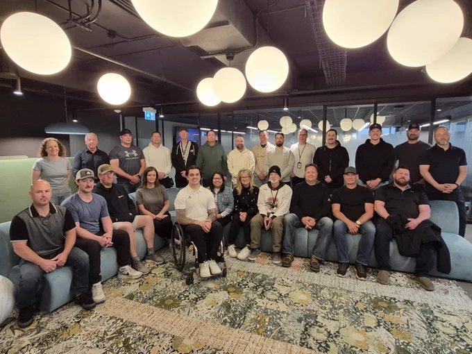 Un groupe de 23 personnes posant ensemble dans un espace de détente de bureau moderne avec des luminaires circulaires au plafond et un tapis à motifs.
 