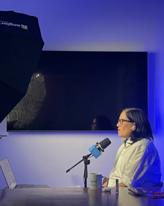 A woman in a white shirt sits at a table with a microphone, participating in a podcast recording session. The mood is professional and focused with a blue-lit background.