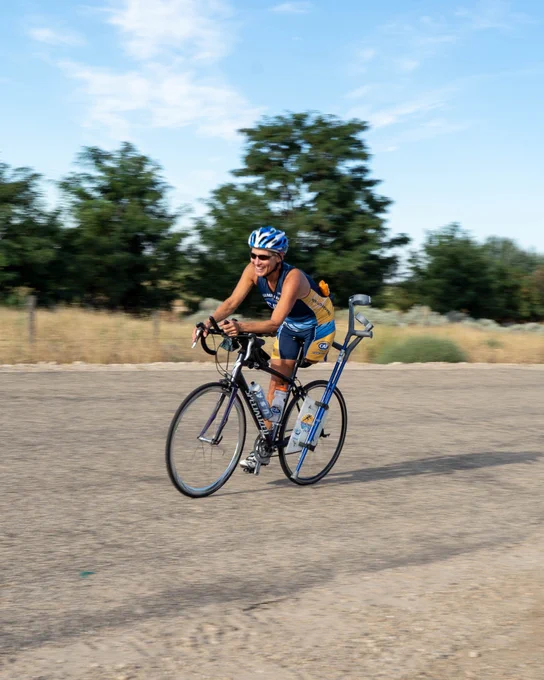 A cyclist in a blue and yellow outfit rides with a crutch attached to their bike on a rural road, with trees in the background.