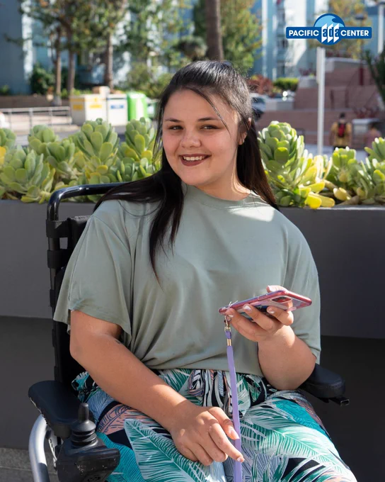 Person sitting in a wheelchair outdoors holding a smartphone with a purple wrist strap, surrounded by green plants and urban buildings.
