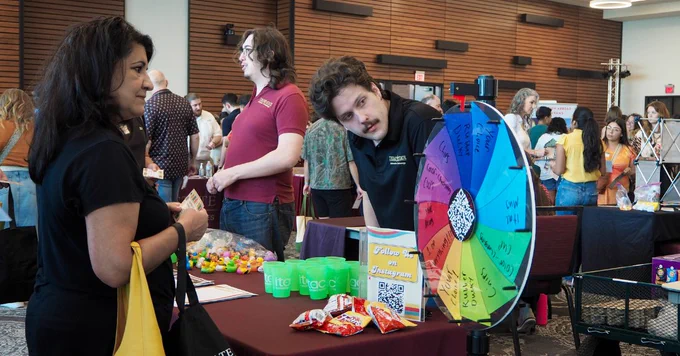 People engaging at a colorful prize wheel booth during a lively indoor event with various tables and attendees in the background.