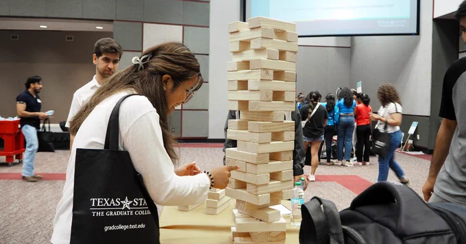 Person playing a large Jenga game indoors with a group of people gathered in the background at an event.