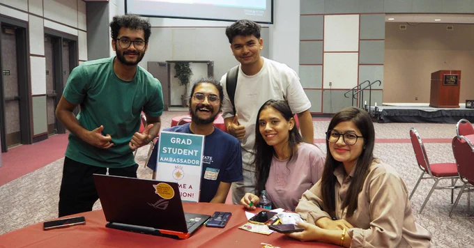 Group of five diverse students gathered around a laptop at a conference table, one holding a sign that reads 'Grad Student Ambassador'.