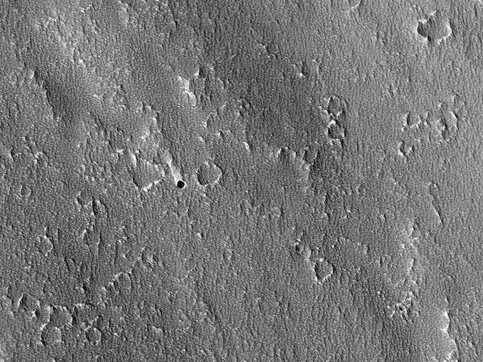 Wider aerial view of flat terrain where the small pit or cave is located on the surface of Mars. Numerous very shallow and infilled impact craters are also visible in the scene. 
