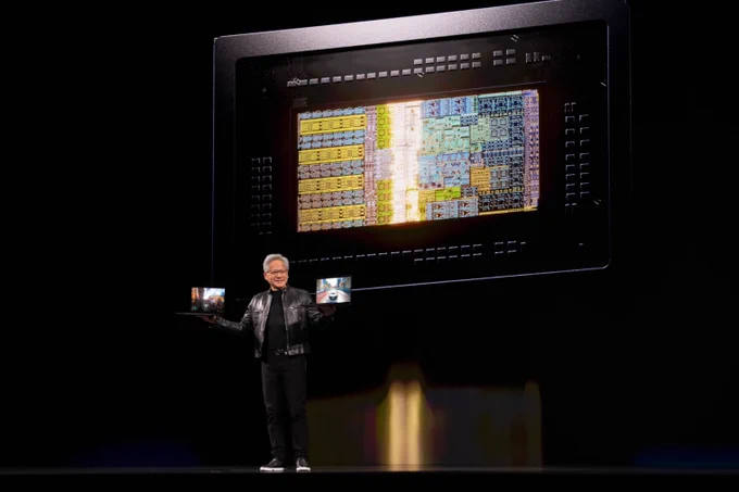 A presenter stands on stage holding two laptop devices, showcasing a large, intricate chip design projected behind him. The chip has various colored circuits and details, illustrating advanced technology.