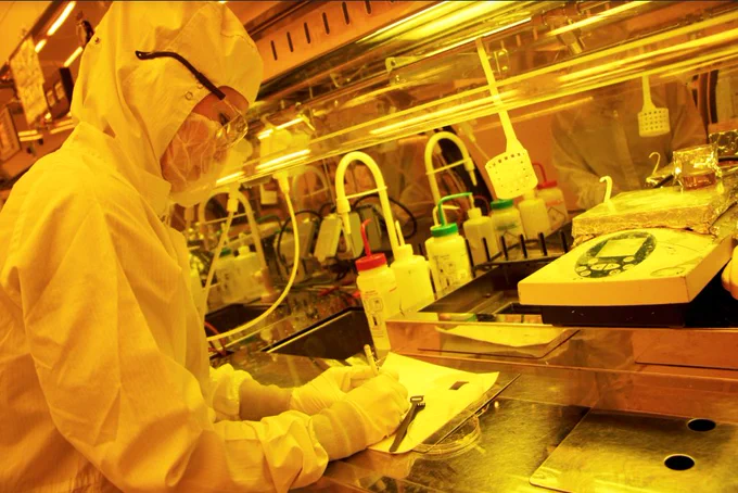 A person in full protective gear takes notes in a lab with chemicals and equipment in front of them. 
